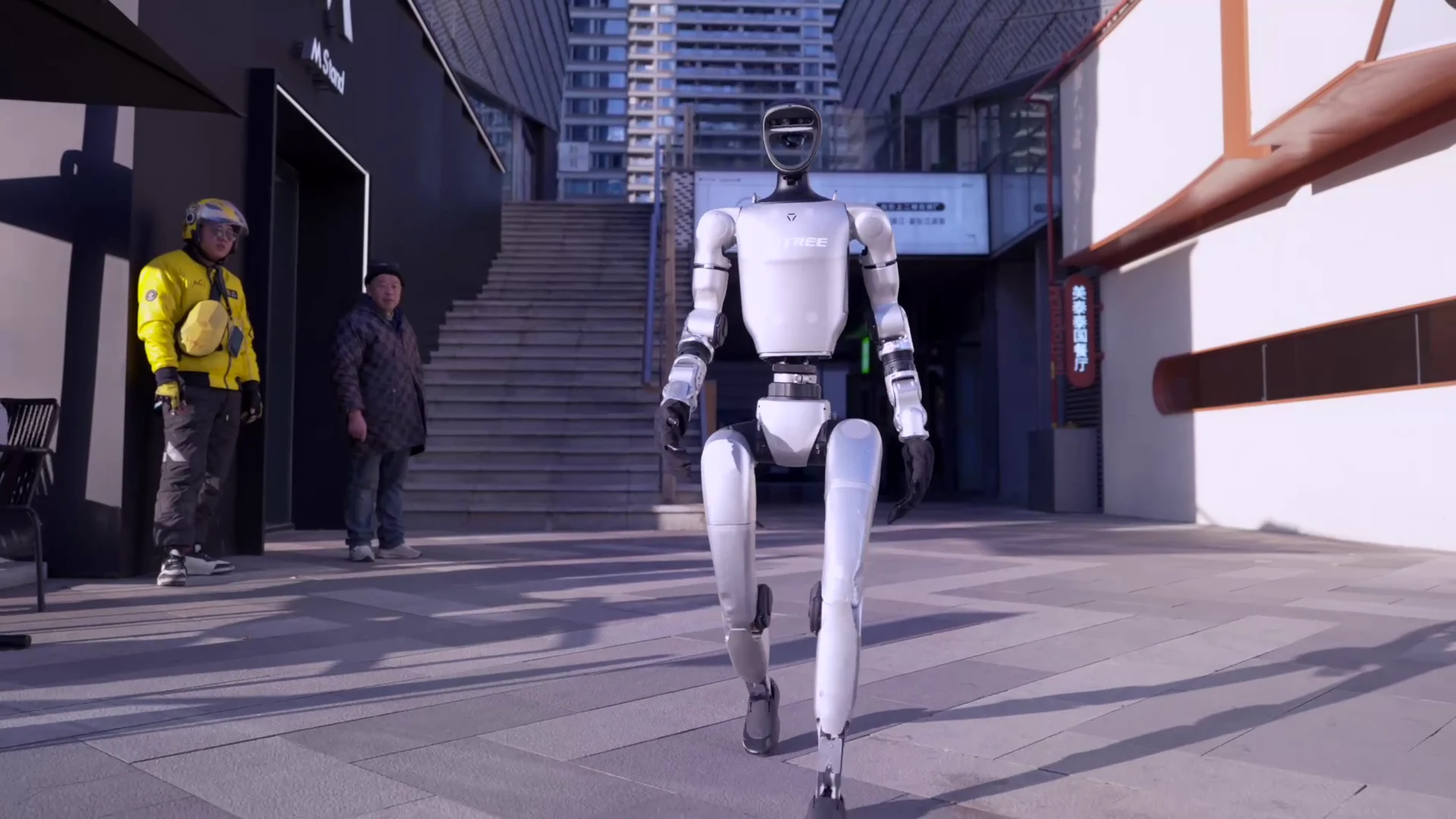 Unitree G1 humanoid robot walking through an urban outdoor plaza while bystanders watch nearby